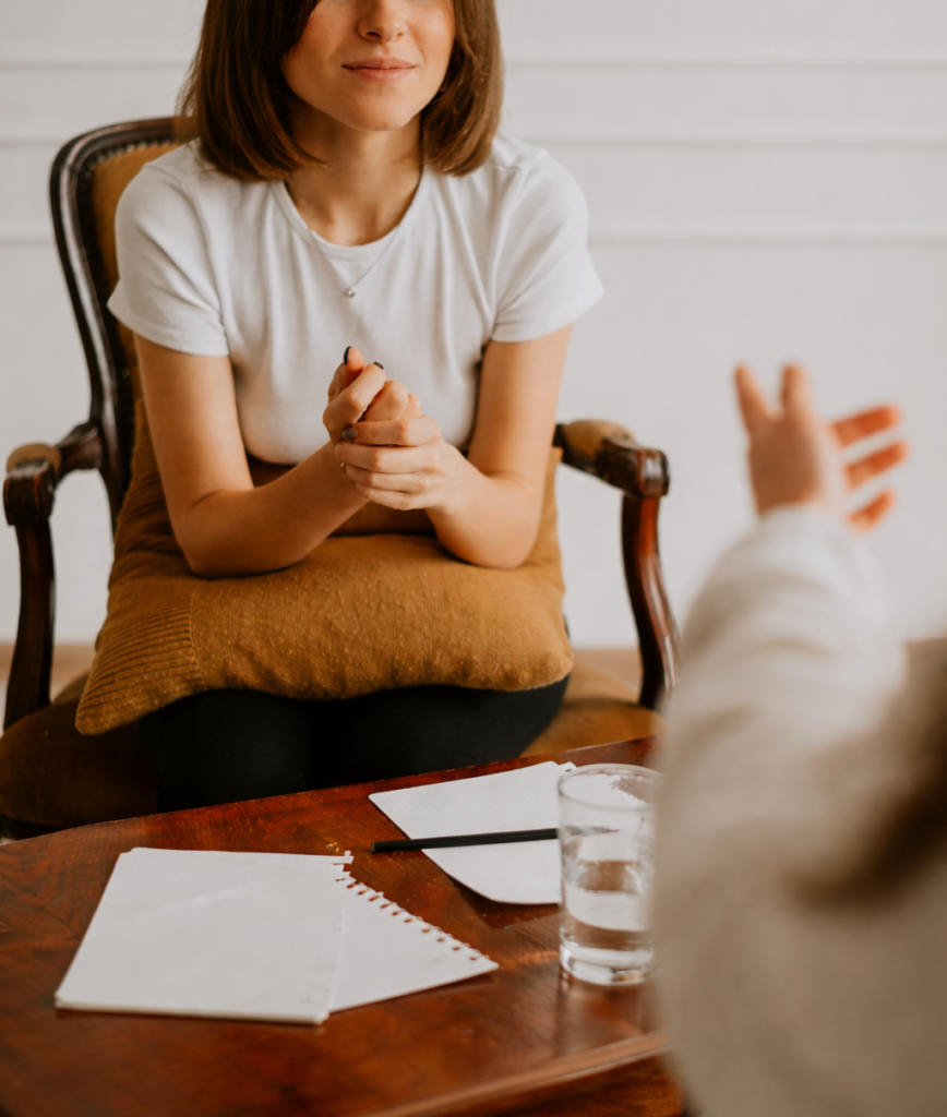 Mulher sentada em uma sessão de terapia, ouvindo atentamente enquanto outra pessoa gesticula ao fundo, em um ambiente acolhedor de escuta e apoio emocional.